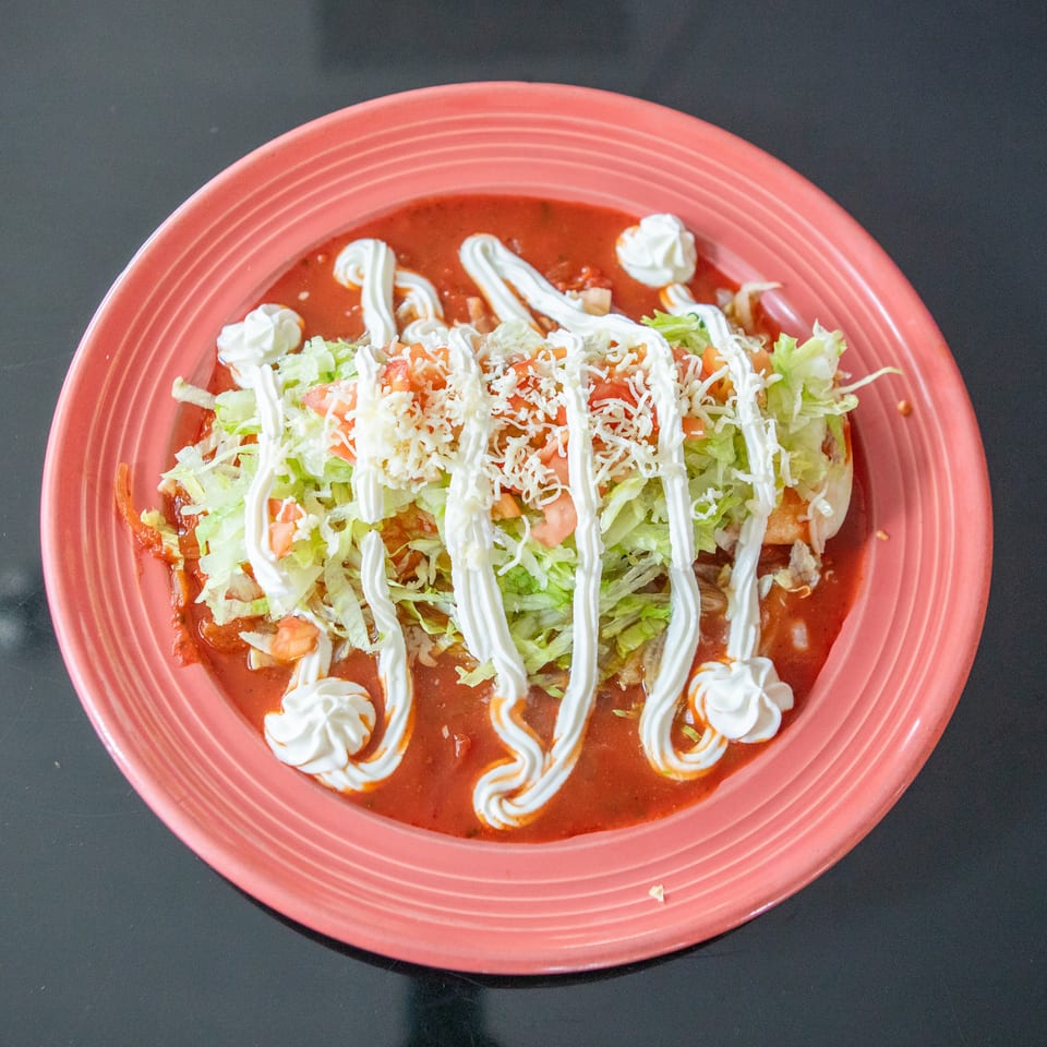Red-sauced Mexican dish topped with shredded lettuce, diced tomatoes, grated cheese, and drizzled crema on a coral plate.