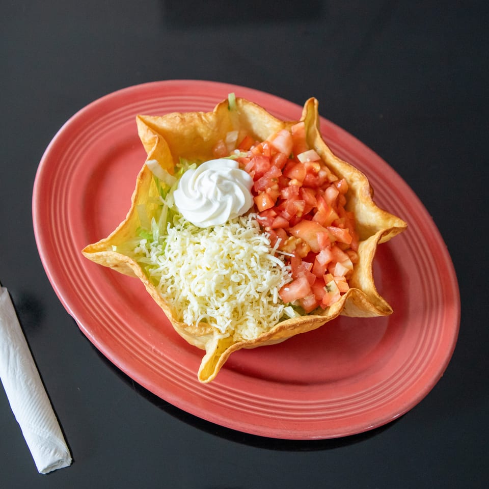 Taco salad in a crisp tortilla bowl topped with lettuce, shredded cheese, diced tomatoes, and sour cream on a red plate.
