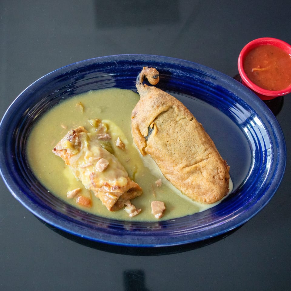 Blue plate holding a cheesy enchilada and crispy folded taco in green sauce, with a side of red salsa.