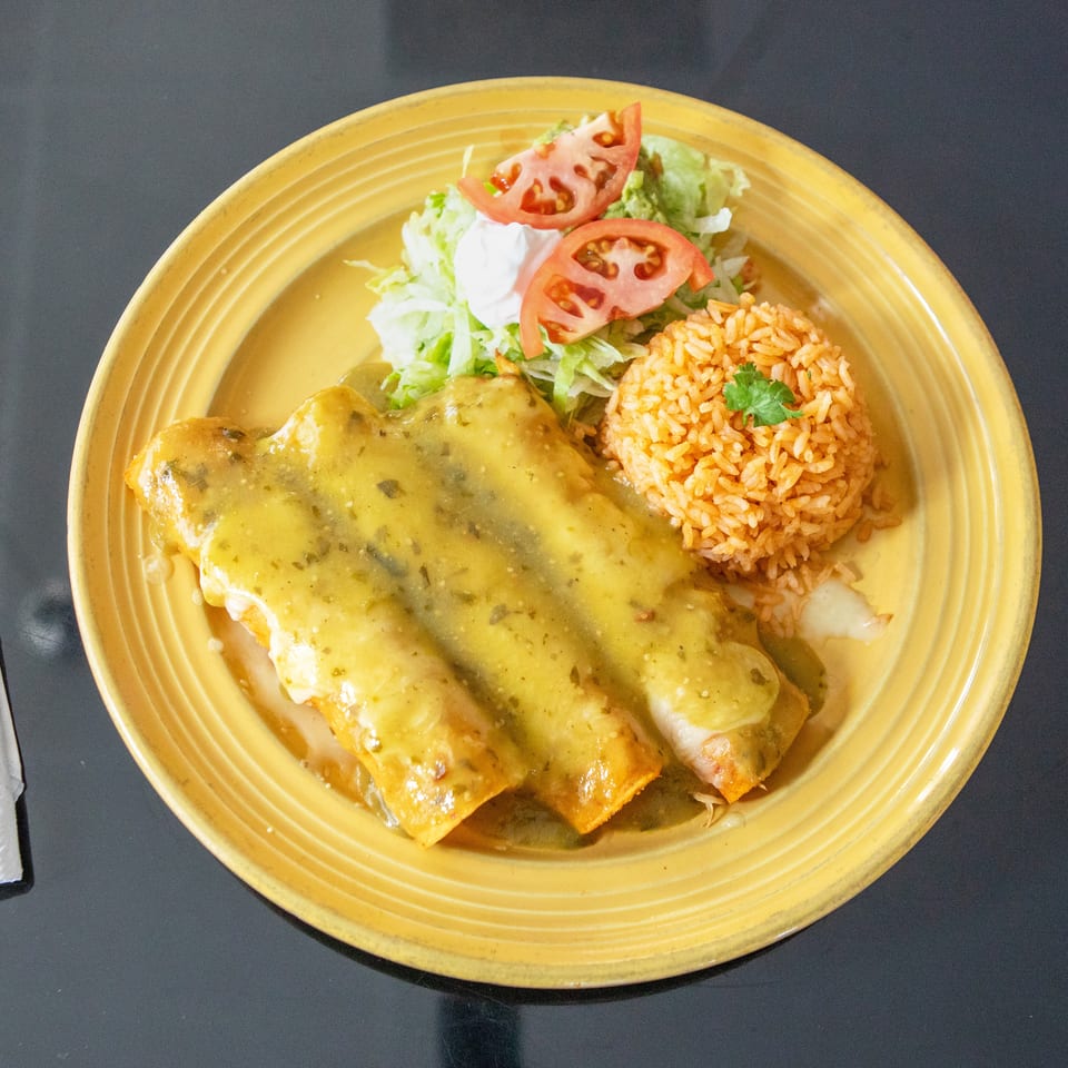 Yellow plate of enchiladas topped with green sauce, served with Mexican rice, lettuce, tomato, and sour cream.